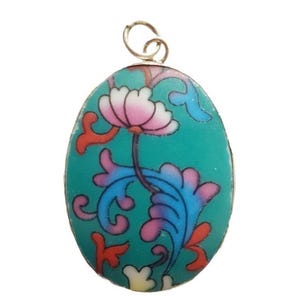 May include: An oval-shaped pendant featuring a vibrant teal background with a decorative floral pattern. The design includes a central pink and white flower with swirling blue, purple, and orange leaf-like accents. A small metal loop is attached at the top.