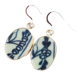 Puede incluir: Un par de pendientes colgantes de forma ovalada con una base de cerámica color crema y un patrón floral azul oscuro. Cada pendiente está enmarcado por un borde de metal dorado y cuelga de un gancho de metal plateado para la oreja.