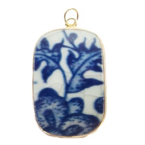 May include: A rectangular pendant featuring a white ceramic base with a blue floral pattern and a visible crackled glaze finish. The piece is encased in a thin gold-toned metal frame and includes a small circular gold-toned metal bail at the top.