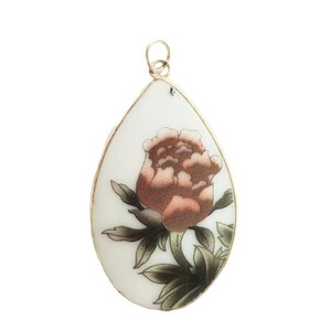 May include: A teardrop-shaped pendant featuring a white ceramic surface with a detailed illustration of a blooming flower in shades of muted pink and brown, accented by dark green leaves, all encased in a thin metallic frame with a top loop.