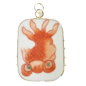 Könnte beinhalten: Ein rechteckiger Anhänger mit einer weißen Keramikoberfläche und einer handgemalten Illustration eines Goldfisches. Der Fisch hat markante Augen mit grünen Zentren und fließende Flossen. Der Anhänger ist von einem dünnen goldfarbenen Metallrand umgeben und verfügt über eine kleine kreisförmige goldfarbene Öse an der Oberseite zur Befestigung an einer Kette oder einem Band.