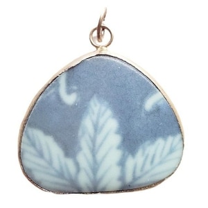 Puede incluir: Colgante con un diseño de hojas en azul claro y blanco. El colgante tiene un marco de metal plateado y un pequeño lazo en la parte superior para una cadena. El diseño recuerda a la porcelana vintage.