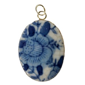 Puede incluir: Un colgante de forma ovalada con un patrón floral azul y blanco sobre una superficie de cerámica con un acabado de esmalte craquelado visible. El colgante está enmarcado por un borde metálico fino e incluye una pequeña anilla metálica superior.