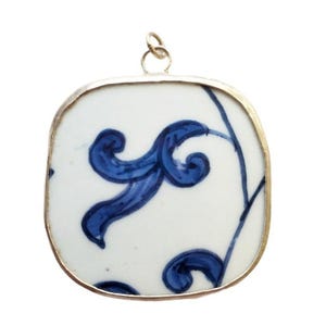 Puede incluir: Un colgante de forma cuadrada con esquinas redondeadas que presenta una base de cerámica blanca decorada con un patrón floral azul pintado a mano. La pieza de cerámica está encerrada en un marco de metal fino de tono dorado e incluye una pequeña anilla metálica circular en la parte superior para sujetarla a una cadena o cordón.