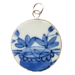 May include: A round, white and blue pendant with a floral design. The pendant has a silver-colored metal frame and a small loop at the top for a chain. The blue floral design is painted on the white background.