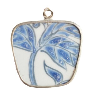 May include: A pendant featuring a piece of white ceramic with a blue painted botanical pattern, encased in a silver-toned metal frame. The ceramic fragment has a rounded, apple-like shape and includes a small metal loop at the top for attachment.