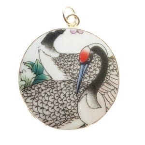 Puede incluir: Colgante redondo con una fianza dorada que presenta un diseño de grulla. La grulla tiene la cabeza roja, el cuello negro y el cuerpo gris estampado. El fondo es blanco con hojas verdes y flores rosas.