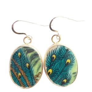 May include: A pair of oval-shaped drop earrings featuring a vibrant peacock feather pattern in shades of teal, yellow, and brown. Each earring is encased in a thin gold-toned metal frame and hangs from a simple gold-toned curved wire hook.