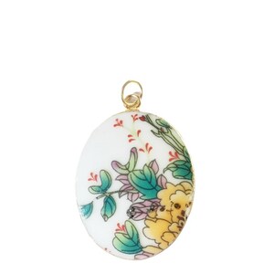 May include: An oval-shaped pendant featuring a white ceramic base with a colorful floral pattern. The design includes yellow and green flowers with thin red accents and green leaves. The pendant is framed in a thin gold-toned metal border with a small loop.
