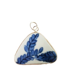 Puede incluir: Un colgante triangular con base de cerámica blanca y un patrón de hojas pintado a mano en azul. La pieza de cerámica está engastada en un fino marco de metal dorado e incluye una pequeña anilla circular de metal dorado en la parte superior.