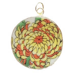 Puede incluir: Colgante redondo con diseño floral. El colgante presenta una flor de crisantemo amarilla y naranja con hojas verdes sobre un fondo blanco. Tiene un lazo de alambre plateado en la parte superior para colgar.