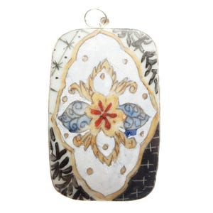 May include: A rectangular pendant featuring a white background with a central yellow and red floral motif surrounded by blue leaf-like shapes. The design is framed by gold-toned lines, with black and white patterned accents in the corners.
