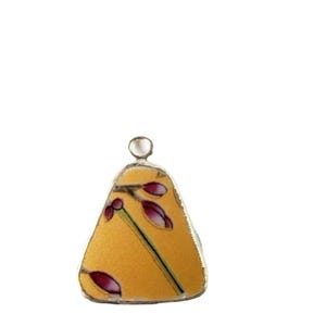 May include: A small, triangular pendant with a gold background and a floral design featuring purple buds and green stems. The pendant has a silver border and a small loop at the top for hanging.