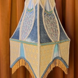 May include: A decorative lampshade with a unique design. The shade features a combination of light blue, beige, and gold fabrics, with intricate patterns and fringe detailing. The lampshade is supported by a clear, decorative base.