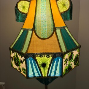 May include: A retro floor lamp with a vibrant, multi-coloured lampshade. The shade is composed of yellow, green, and blue panels, trimmed with dark green edging and fringe. Floral patterns and decorative elements enhance the vintage aesthetic.
