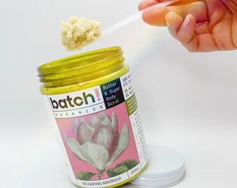 May include: A jar of "Batch Botanics" butter and sugar body scrub, with a magnolia flower illustration. The jar is yellow with a white lid. A spoonful of the scrub is held above the jar. The label reads "Enchanting Magnolia" and the jar contains 300ml.