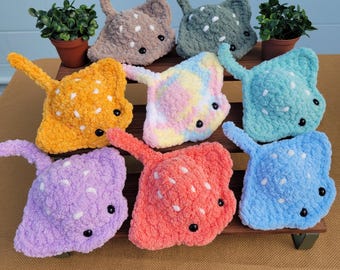 May include: A collection of handmade crochet stingrays in various colors, including yellow, pink, blue, and purple. Each plush stingray features white spots and black bead eyes. The toys are arranged on a wooden display.