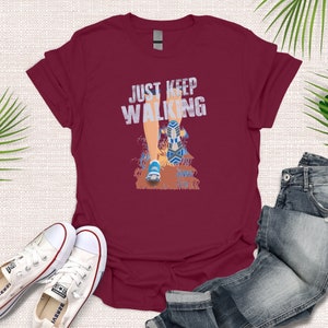 May include: Burgundy t-shirt with a graphic of a person's leg walking with the text "JUST KEEP WALKING" printed on the front.