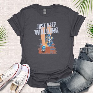 May include: A dark gray t-shirt with the text "JUST KEEP WALKING" in white lettering. A cartoon illustration of a person's leg walking is below the text.