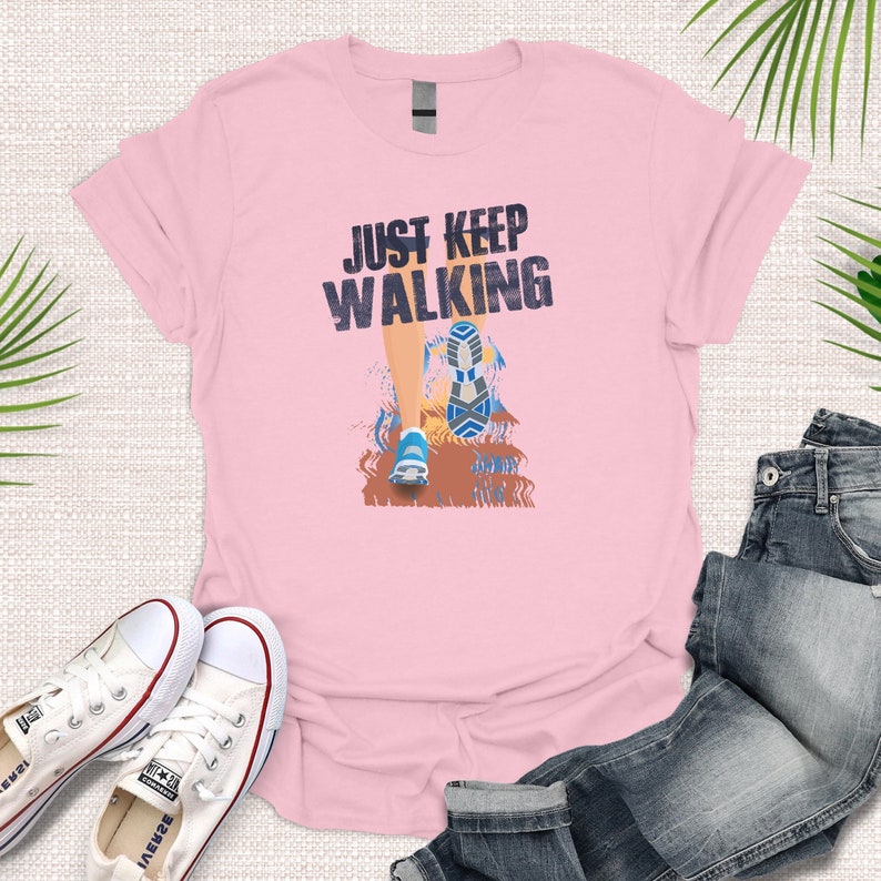 May include: Pink t-shirt with a graphic of a person walking with the text "JUST KEEP WALKING" printed on it.
