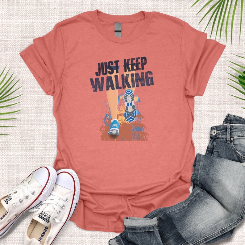 May include: A coral colored t-shirt with the text "JUST KEEP WALKING" and a graphic of a person's legs and feet walking. The person is wearing blue and white sneakers.