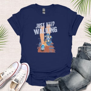 May include: Navy blue t-shirt with a graphic of a person's leg walking with the text "JUST KEEP WALKING" in white.