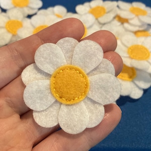 May include: White felt daisy flower with a yellow center. The daisy has white petals and a yellow center with a stitched border. Several other daisies are visible in the background against a blue surface.