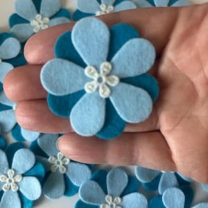 May include: A collection of layered felt flower cutouts in shades of light and dark blue. Each flower features six rounded petals in a light blue hue, layered over six dark blue petals, and topped with a small, white, six-petaled centre detail.
