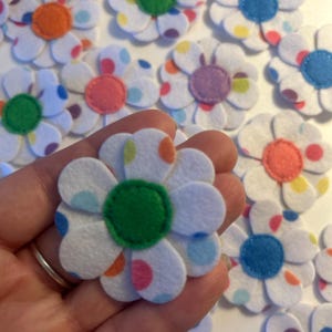 May include: A collection of white felt flower cutouts featuring multi-coloured polka dots on the petals and solid-coloured circular centres in shades of green, orange, blue, pink, and purple, arranged in a scattered pattern on a white surface.