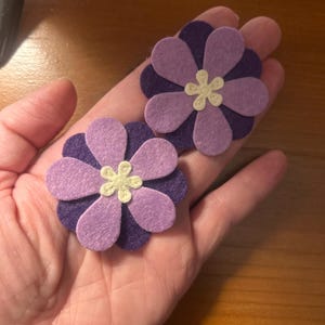 May include: Two layered felt flower appliques featuring dark purple bottom petals and lighter lavender top petals. Each flower has a small, cream-coloured embroidered centre detail in the shape of a six-petaled blossom, crafted from soft felt material.