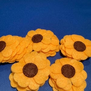 May include: Six felt sunflowers with yellow petals and brown centres are arranged on a blue background. The handmade flowers have a soft texture. Each flower is approximately 10 cm in diameter.