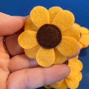 May include: A handcrafted felt flower applique featuring bright yellow rounded petals arranged in a circular pattern around a dark brown circular center. The flower is constructed from layered felt material with visible stitching securing the center piece.