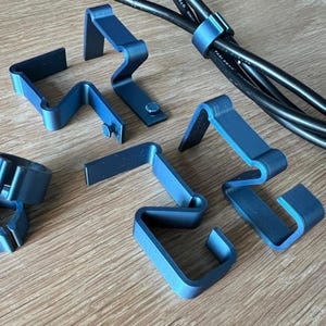 Peut inclure: Un assortiment d'attaches et de supports de gestion de câbles en métal bleu. Les clips sont de formes variées, dont un support circulaire et plusieurs clips en forme de Z. Des câbles noirs sont fixés avec un clip bleu.