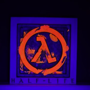 Könnte beinhalten: Ein quadratisches, gerahmtes Kunstwerk mit dem orangefarbenen Half-Life-Logo, einem Lambda-Symbol in einem Kreis, vor einem strukturierten, dunkelblauen Hintergrund. Die Worte "HALF-LIFE" sind unten in Weiß gedruckt. Das Kunstwerk wird von blauem Licht beleuchtet.