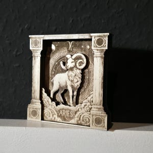 Può includere: Scultura in legno del segno zodiacale dell'Ariete. L'opera presenta un ariete in piedi sulle nuvole, incorniciato da colonne. La parola "ARIES" è in basso. Lo sfondo ha un design celeste con stelle e il simbolo dell'Ariete.