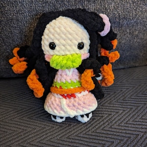 May include: A crocheted plush toy of a character with black hair, pink and white clothing, and a green face mask. The toy has large black eyes and orange hair accents.