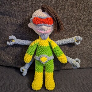 May include: A handmade crochet doll with a superhero theme. The doll features a white head with brown hair, red and blue eye mask, and a green and yellow body. It has four blue arms and yellow feet. The doll is sitting on a gray surface.