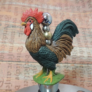 May include: A rooster-shaped bottle stopper with a green, brown, and red design. The stopper is made of metal and has a silver base. The rooster is perched on a green base with yellow feet.