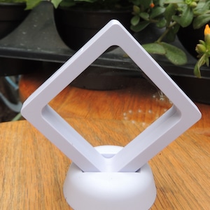 May include: White plastic display stand with a square frame, measuring 3.5 inches. The stand is designed to hold small items, such as jewelry or coins.