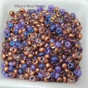 May include: A close-up of a bowl filled with a variety of small, round beads. The beads are in shades of purple, blue, and copper. Some of the beads have a metallic finish, while others are iridescent.