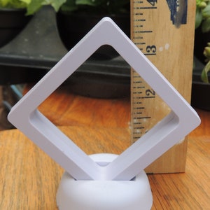 May include: White plastic display stand with a square opening, measuring 3.5 inches. The stand is designed to hold and display small items, such as jewelry or coins. The stand is on a wooden surface.