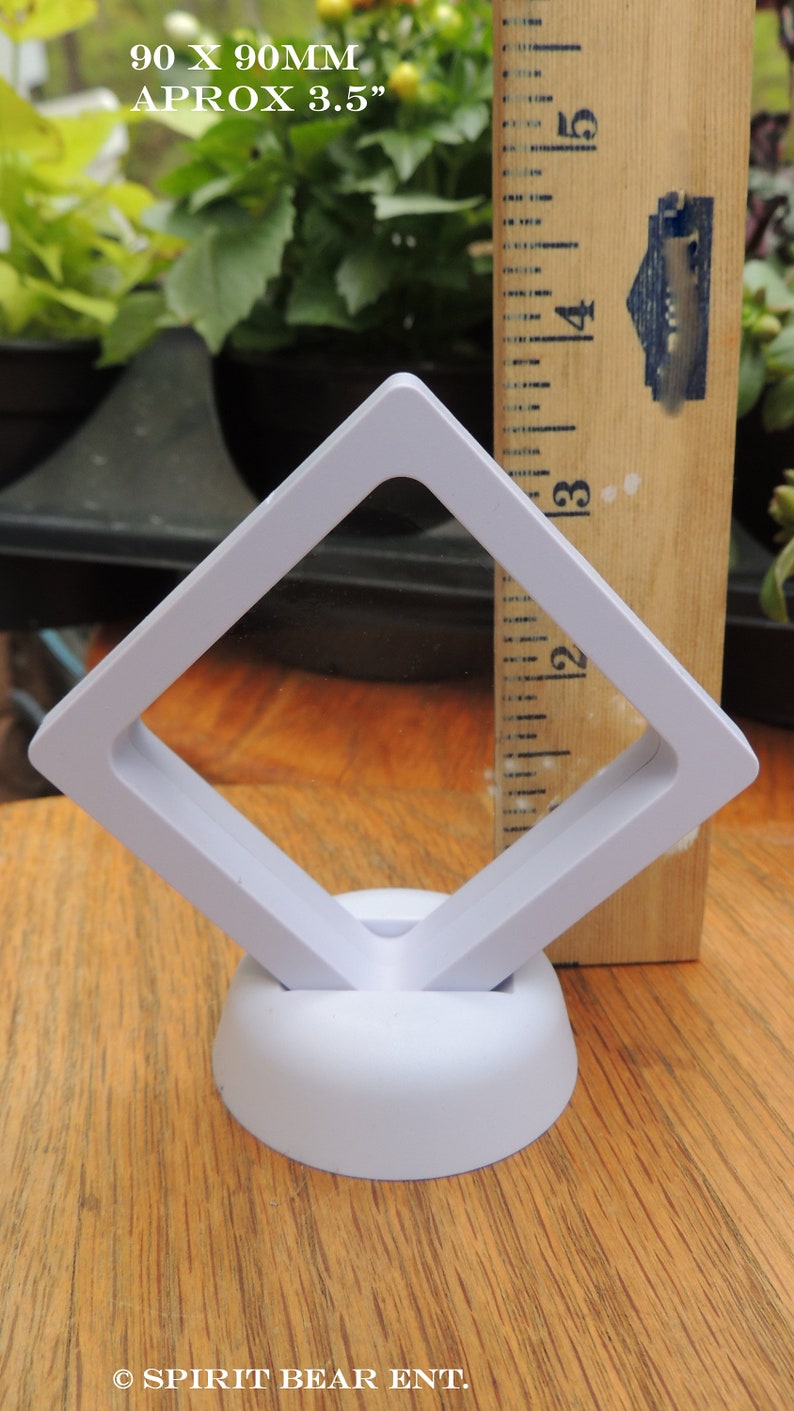 May include: White plastic display stand with a square opening, measuring 3.5 inches. The stand is designed to hold and display small items, such as jewelry or coins. The stand is on a wooden surface.