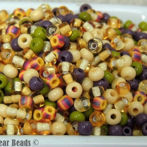 May include: A close-up of a bowl filled with a variety of colourful beads. The beads are in shades of cream, green, purple, orange, and yellow. Some of the beads are transparent, while others are opaque. The beads are arranged in a random pattern.