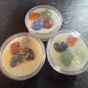 May include: Three clear circular plastic containers, each holding a collection of small, polished stone carvings shaped like elephants. The stones feature various colors including orange, green, blue, brown, gray, and tan, resting on white foam.