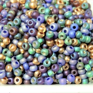 May include: A collection of iridescent glass seed beads in shades of blue, green, and gold. The beads are small and round, and they have a slightly metallic sheen.