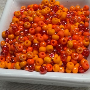 May include: A white square dish filled with a variety of orange and red glass beads. The beads are in various shades of orange, from light to dark, with some translucent red beads mixed in. These beads are ideal for jewelry making and crafts.