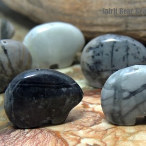 May include: A collection of six small, hand-carved stone bear figurines. The bears are made from different types of stone, including black, white, and gray. Each bear has a unique pattern and texture. Spirit Bear Beads © 2013.