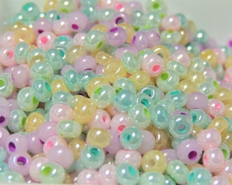 May include: A close-up shot of a pile of iridescent beads in pastel colors. The beads are a mix of pink, yellow, green, and purple, with a pearlescent sheen. Each bead has a hole running through the center, and they are scattered randomly.