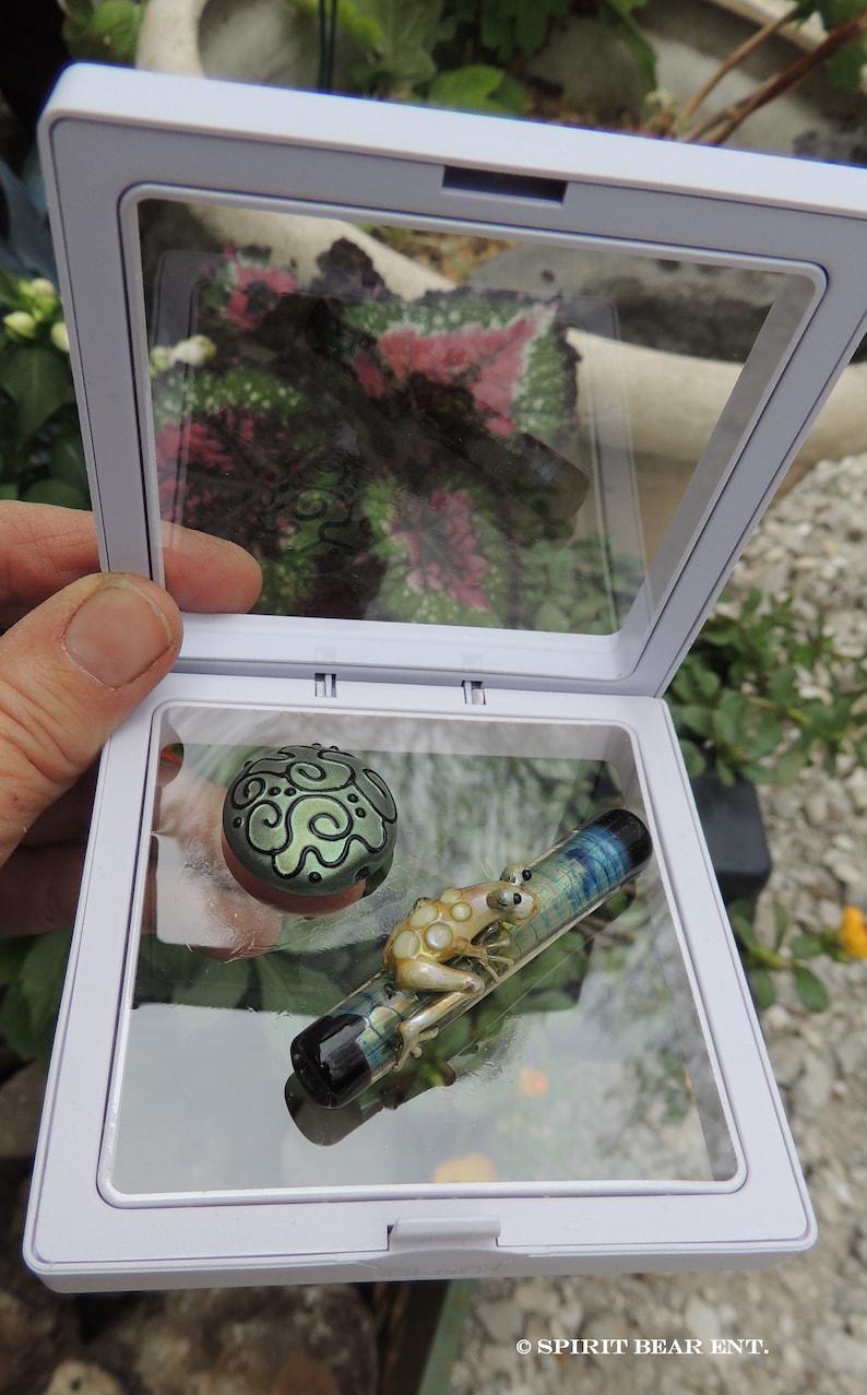 May include: A white display case with a glass top, showcasing a green and black glass bead with a swirling pattern and a glass frog figurine on a blue and green glass rod.