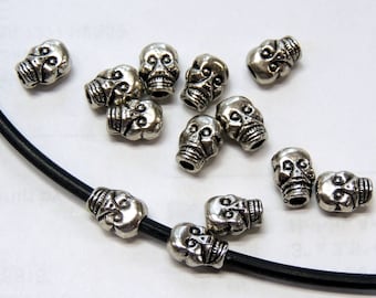 May include: A close-up of silver skull beads, some of which are strung on a black cord. The beads are about 1 cm in diameter and have a detailed skull design.